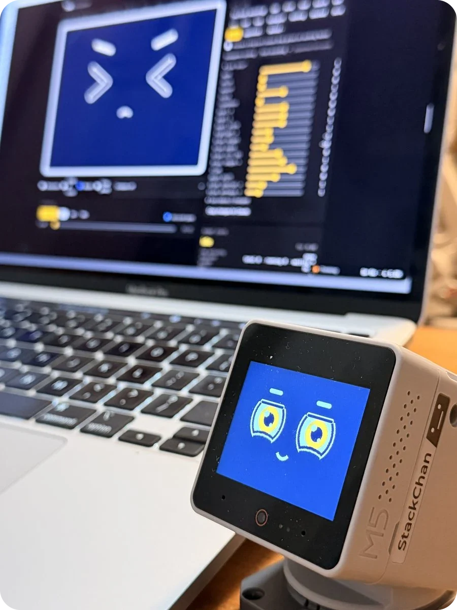Toki's first face on the robot, cream outlines on dark navy with ringed yellow eyes, in front of a laptop running the face simulator