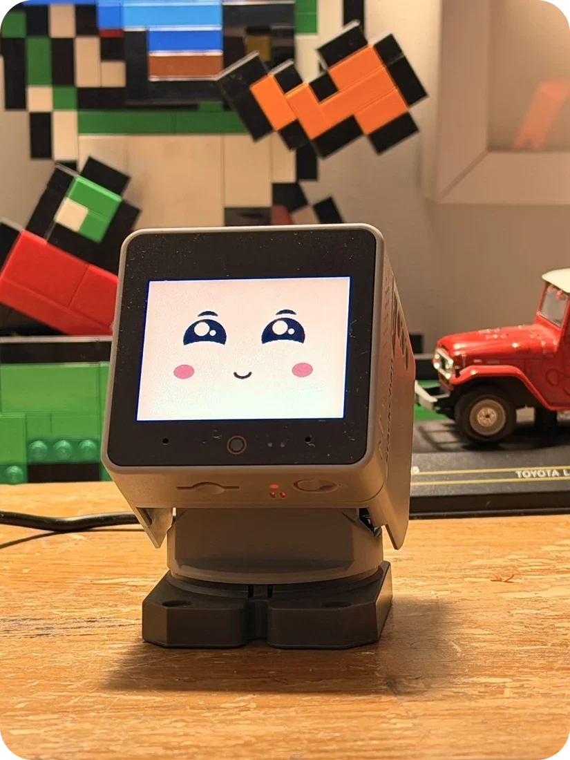 Toki on a wooden desk, a small square robot with a cream screen showing a happy face with big shiny eyes and pink cheeks